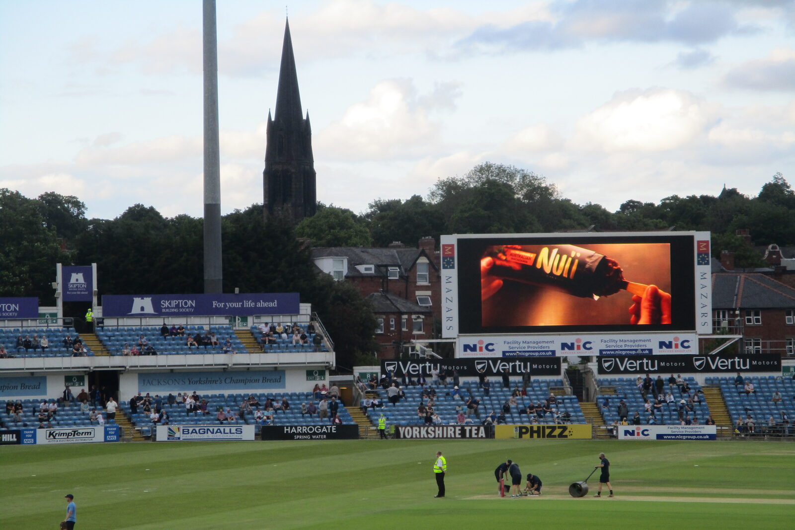 Two open tiers of blue seats in Headingley’s North East Stand beside the scoreboard and church spire.